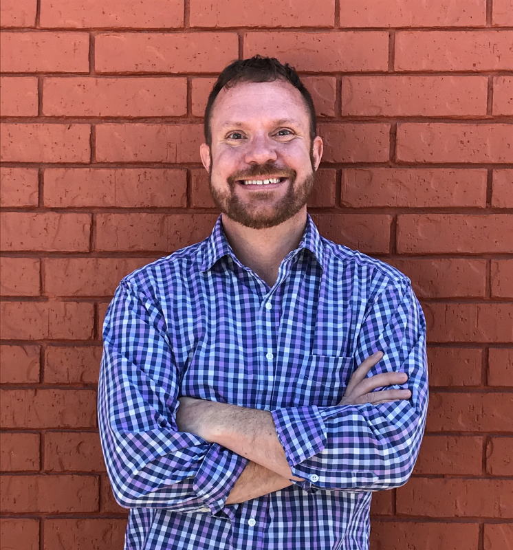 Head shot of Reverend Steven Gaines, white male with beard, smiling with arms crossed, wearing a blue check shirt, in front of a red brick wall.